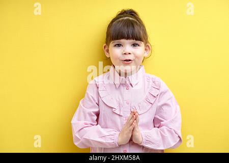 petite fille mendiant à acheter, demandez quelque chose. enfant doux dans le chemisier rose garder les mains ensemble, isolé sur fond jaune, regarder l'appareil photo Banque D'Images