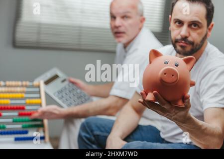 Homme déçu montrant une banque de pigeons tandis que son père âgé calcule ses économies avec une calculatrice et un comptoir à la maison Banque D'Images
