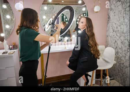 Maquilleur qui photographie un client dans un salon de beauté. Jeune femme aux cheveux longs assise sur une chaise et regardant un spécialiste de la beauté pendant que la femme prend des photos avec un smartphone. Banque D'Images