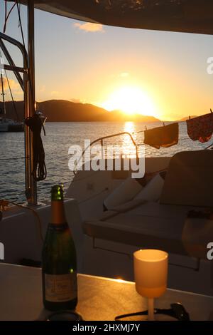 Bouteille de champagne et verre silhouetés contre le coucher du soleil sur un yacht avec deux paires de boxers de natation séchant sur les fils de sécurité du yacht Banque D'Images