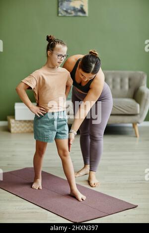 Portrait vertical pleine longueur de la jeune fille avec le syndrome de Down s'exerçant à l'intérieur avec une femme instructeur aidant Banque D'Images