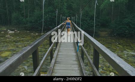 Femme marchant le long du pont suspendu seul dans un cadre pittoresque de la Forêt verte. Mouvement lent de la femme à poil sombre marcher sur le pont suspendu dans la forêt épaisse verte. Aucune autre personne n'est dans la photo. Banque D'Images