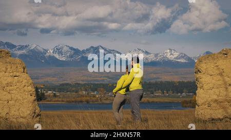 Homme et femme dans le vert jaune sportswear. Couple de voyageurs adorables embrassent et embrassent près de la vieille pierre en profitant du paysage des montagnes. Deux voyageurs se promo dans les montagnes enneigées. Banque D'Images