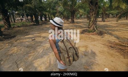 Femme touriste avec la tresse marche autour de la croissance de jeunes arbres avec des feuilles luxuriantes à la ferme de palmiers à huile elaeis guineensis le jour ensoleillé. Concept de culture exotique, voyage dans les pays tropicaux Banque D'Images