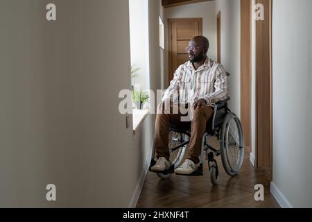 Jeune Afro-américain handicapé regardant par la fenêtre tout en étant assis sur un fauteuil roulant dans le couloir de l'appartement Banque D'Images