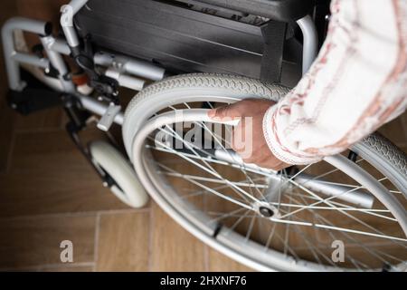 Vue au-dessus de la main d'un jeune homme handicapé tournant la roue du fauteuil roulant tout en se déplaçant vers le bas du plancher en bois Banque D'Images