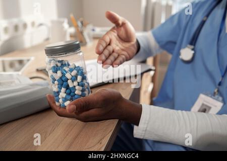 Mains de jeunes hommes afro-américains cliniciens tenant un pot avec des pilules blanches et bleues tout en les recommandant au patient Banque D'Images