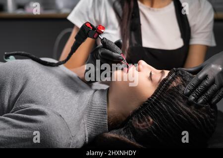 femme ayant le maquillage permanent sur ses lèvres dans le salon d'esthéticienne. Le maître applique le pigment à la face. Banque D'Images