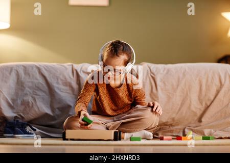 Portrait d'une jeune fille avec le syndrome de Down jouant avec des blocs de jouets seuls et portant des écouteurs antibruit, concept de surstimulation, espace de copie Banque D'Images