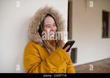 Jeune femme heureuse avec le syndrome de Down weraing parka, se penchant le mur et en utilisant le smartphone. Banque D'Images