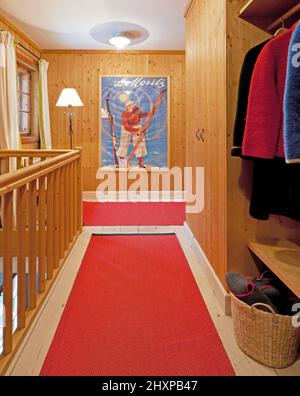 Hall d'entrée dans une maison de style alpin avec murs lambrissés, rambardes en bois, moquette rouge, armoire avec blousons, lampes et affiche bleue Banque D'Images