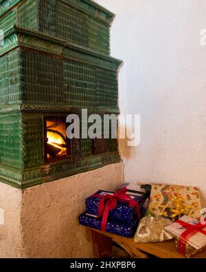 Vieux poêle en faïence de vert avec feu vu dans la porte ouverte et des colis de Noël disposés près de lui Banque D'Images