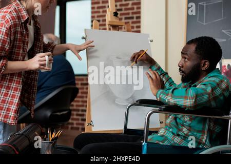 Professeur peintre montrant la technique de dessin à un étudiant handicapé travaillant au projet de peinture pendant la leçon artistique en studio de créativité. Divrse le modèle d'esquisse d'équipe à l'aide d'un crayon graphique Banque D'Images