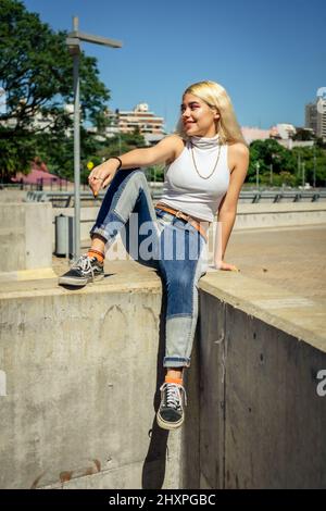 Un portrait vertical d'une belle jeune femme latine assise souriante sur une rampe de ciment avec une jambe pendante un jour ensoleillé. Banque D'Images