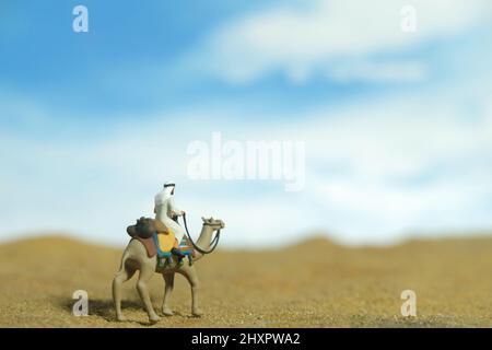 Photographie miniature de personnage de jouet de personnes. Un homme portant des vêtements kandura blancs à cheval à dos de chameau dans un désert dans un ciel lumineux. Photo d'image Banque D'Images