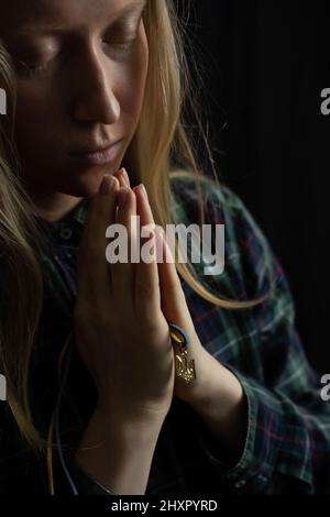 La femme blonde ukrainienne prie pour l'Ukraine pendant la guerre avec l'emblème national Trident dans les palmiers de près. La Russie a attaqué l'Ukraine le 24 février 2022 Banque D'Images