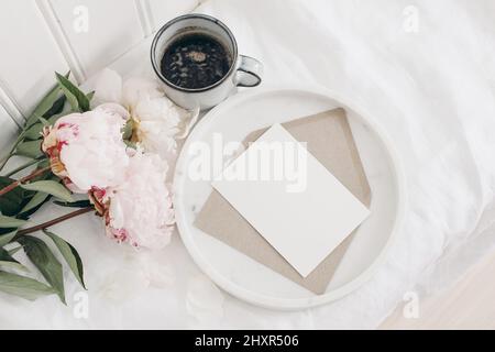 Fleurs de pivoines roses en fleurs, bouquet allongé sur une table. Mur en bois blanc. Carte de vœux vierge maquette sur plateau en marbre. Tasse de café, petit déjeuner au lit Banque D'Images