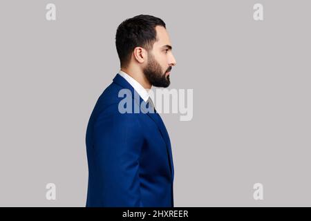 Portrait en vue latérale d'un homme à barbe plein de confiance regardant un appareil photo avec une expression sérieuse, homme d'affaires déterminé sans sourire, portant un costume de style officiel. Prise de vue en studio isolée sur fond gris. Banque D'Images