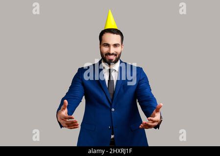 Entrez dans mes bras. Sympathique homme souriant dans un cône de fête donnant des câlins aux mains tendues, accueillant et accueillant, portant un costume de style officiel. Prise de vue en studio isolée sur fond gris. Banque D'Images