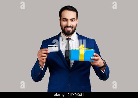 Portrait d'un homme barbu souriant et positif montrant une carte de crédit et une boîte cadeau, regardant avec étonnement, banque, portant un costume de style officiel. Prise de vue en studio isolée sur fond gris. Banque D'Images