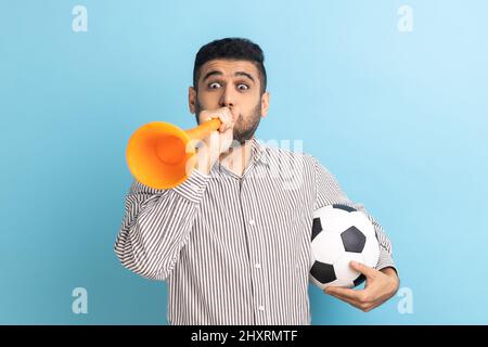 Homme d'affaires extrêmement positif satisfait avec la corne de taureau tenant le ballon de football, célébrant la victoire de l'équipe de football préférée, portant une chemise rayée. Studio d'intérieur isolé sur fond bleu. Banque D'Images