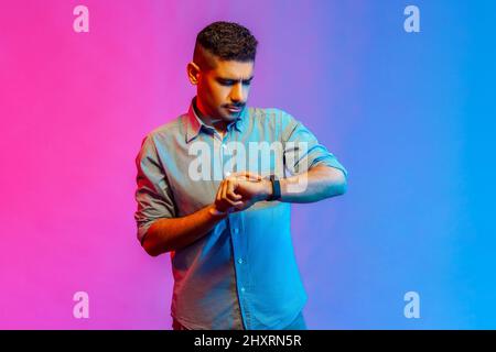 Portrait d'un jeune adulte concentré homme dans une chemise debout regardant sa montre intelligente, vérifier l'heure, étant pressé de se rencontrer. Studio d'intérieur isolé sur fond de néon coloré. Banque D'Images