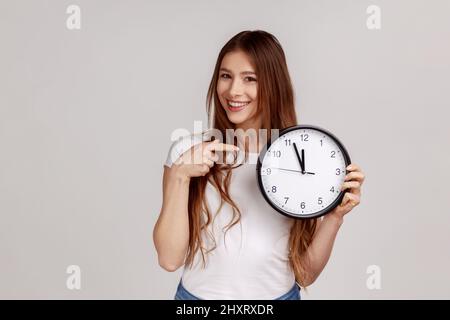 Portrait d'une femme heureuse et attrayante tenant une grande horloge murale pointant, regardant l'appareil photo avec un sourire agréable, le temps d'aller, portant un T-shirt blanc. Prise de vue en studio isolée sur fond gris. Banque D'Images