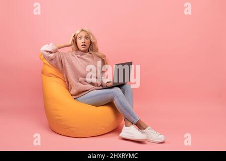 Concept d'échéance. Une jeune femme choquée assise dans un fauteuil avec un ordinateur portable et saisissant sa tête, fond rose Banque D'Images