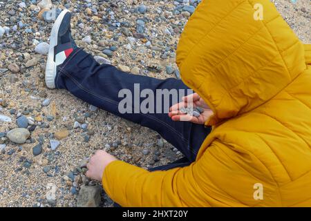 Homme au manteau jaune, assis sur la plage de ramasser des cailloux et des coquillages Banque D'Images