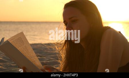 Une jeune femme lit un livre en étant couché sur la plage au coucher du soleil Banque D'Images