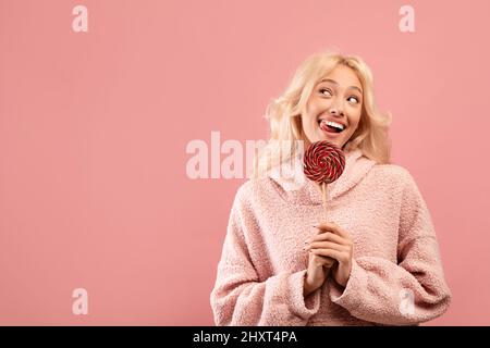 Concept de dent douce. Une dame enjouée tient des bonbons sur le bâton, regardant de côté l'espace libre et souriant, fond rose Banque D'Images