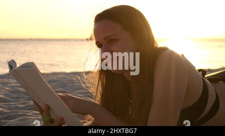 Une jeune femme lit un livre en étant couché sur la plage au coucher du soleil Banque D'Images