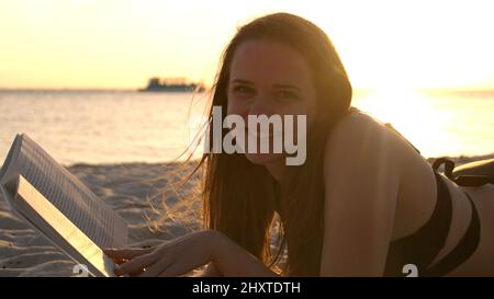 Une jeune femme lit un livre en étant couché sur la plage au coucher du soleil Banque D'Images