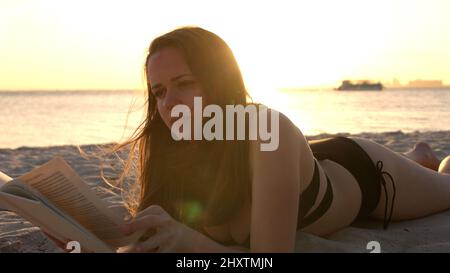 Une jeune femme lit un livre en étant couché sur la plage au coucher du soleil Banque D'Images