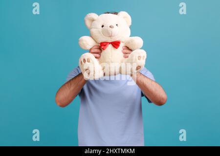 Portrait d'un homme anonyme inconnu barbu cachant son visage derrière un ours en peluche blanc, surprise romantique pour sa girflriend. Studio d'intérieur isolé sur fond bleu. Banque D'Images
