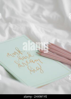 Un carnet avec "Work Hard Dream Big" écrit dessus et des crayons roses à côté, sur une feuille blanche Banque D'Images