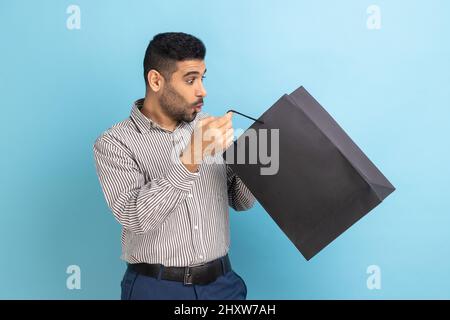 Surprise homme d'affaires barbu regardant à l'intérieur de sac de shopping avec les yeux grands ouverts la bouche dans étonnement, choqué par l'achat, portant une chemise rayée. Studio d'intérieur isolé sur fond bleu. Banque D'Images