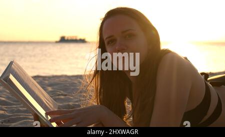 Une jeune femme lit un livre en étant couché sur la plage au coucher du soleil Banque D'Images