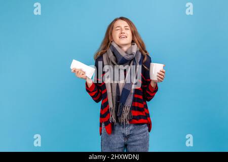 Femme dépairée portant un chandail rayé de style décontracté, enveloppé dans un foulard tenant le mouchoir et la tasse à thé, criant avec la colère de haine, la maladie de la grippe. Studio d'intérieur isolé sur fond bleu. Banque D'Images