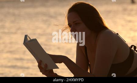 Une jeune femme lit un livre en étant couché sur la plage au coucher du soleil Banque D'Images