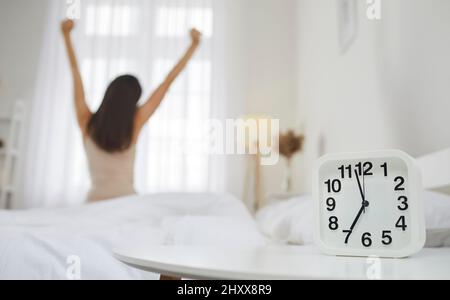 Gros plan du réveil sur fond duquel une femme heureuse assise sur le lit s'étire après s'être réveillée. Banque D'Images