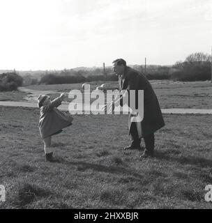 1950s, historique, à l'extérieur sur un morceau de prairie ouverte, un père dans un pardessus, jouant un jeu de capture avec une petite balle avec sa jeune fille, qui est dans un petit manteau et portant un chapeau de carcajou, Angleterre. ROYAUME-UNI. Prendre ou jouer la capture, avec un objet, est une activité amusante pour un parent et un enfant à faire ensemble. Une voiture de l'époque est garée sur un chemin. Banque D'Images