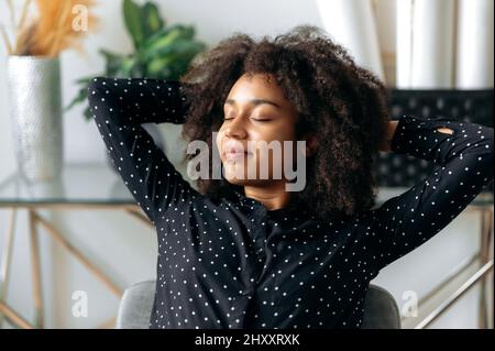 Gros plan d'une charmante femme, directrice, directrice financière, calme et à la peau sombre, assise dans une posture détendue au bureau, les yeux fermés, les mains derrière la tête, se reposant, souriant Banque D'Images