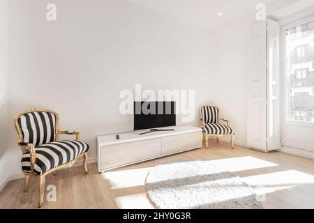 Séjour avec grand buffet en bois, table tv, canapé bleu, grande fenêtre avec portes et volets en bois donnant sur un balcon et fauteuil or wi Banque D'Images