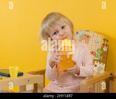 Une jeune fille gaie est assise à la maison dans un fauteuil pour enfants et de boire du jus d'une bouteille de non-arrosage. Arrière-plan jaune Banque D'Images