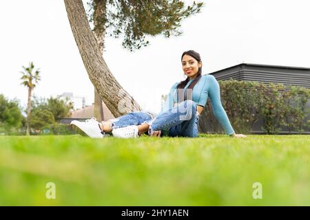 Au niveau du sol, une femme indienne positive regarde la caméra tout en étant assise sur une pelouse verte en ville le jour de l'été Banque D'Images