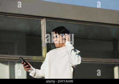 Un homme indien positif écoute de la musique dans un casque sans fil tout en surfant sur un téléphone portable moderne dans une rue ensoleillée près du bâtiment de la ville Banque D'Images
