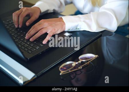 Rognez une femme anonyme qui navigue sur un netbook tout en travaillant à table avec des lunettes dans un bureau moderne et léger pendant la journée de travail Banque D'Images