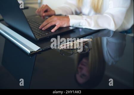 Rognez une femme anonyme qui navigue sur un netbook tout en travaillant à table avec des lunettes dans un bureau moderne et léger pendant la journée de travail Banque D'Images