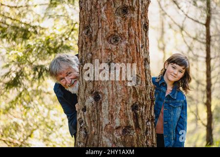 Grand-père âgé positif avec cheveux gris et barbe avec petite fille souriante dans la veste en denim se cachant derrière l'arbre et regardant l'appareil photo dans la forêt sur b Banque D'Images